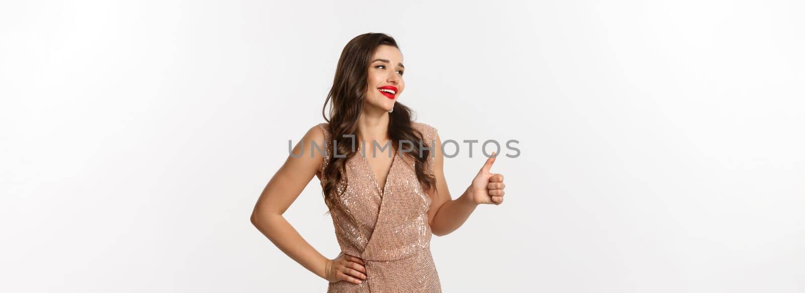 Concept of celebration, holidays and party. Beautiful female model in luxurious dress, looking left and showing thumb-up in approval, praise good choice, standing over white background.