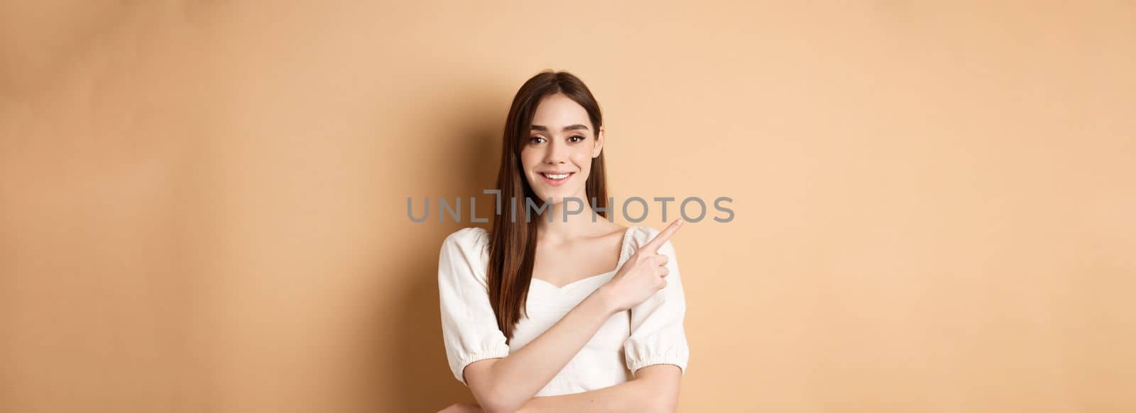 Young cheerful female model in romantic dress pointing finger at upper left corner, smiling pleased at camera, showing her choice, recommending product, beige background by Benzoix