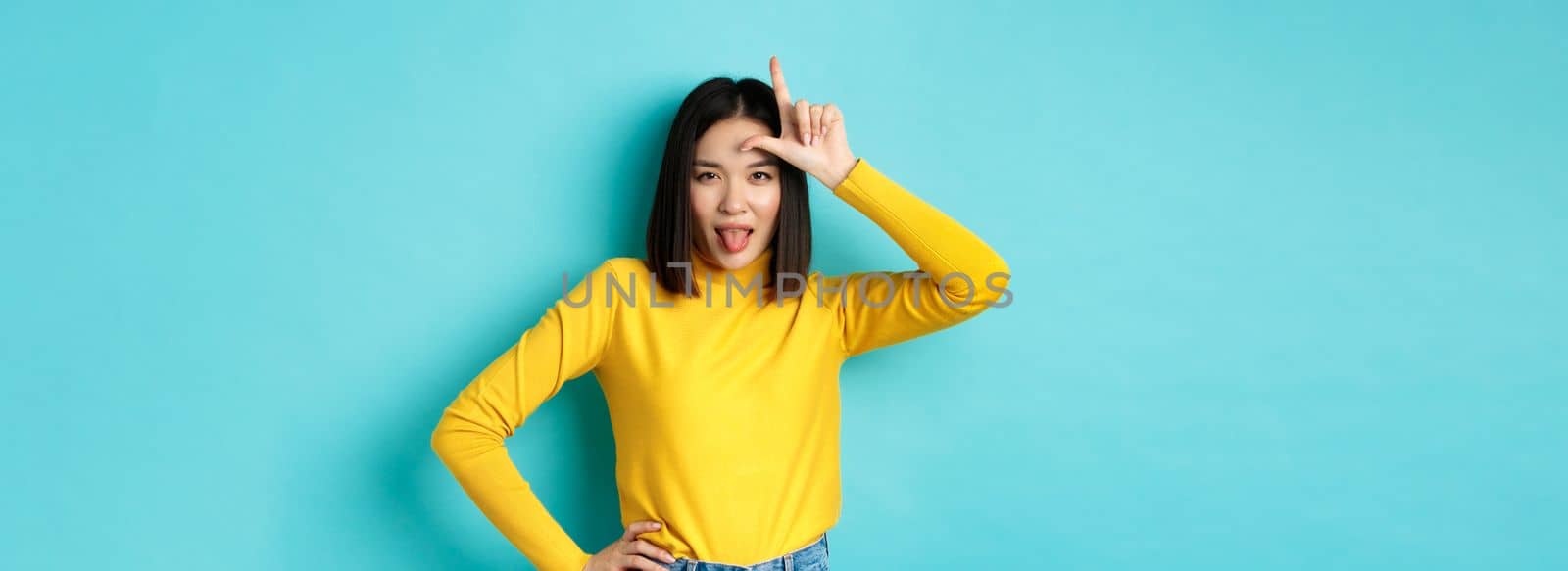 Asian girl winner mocking over lost team, showing loser sign on forehead and sticking tongue ignorant, standing over blue background by Benzoix