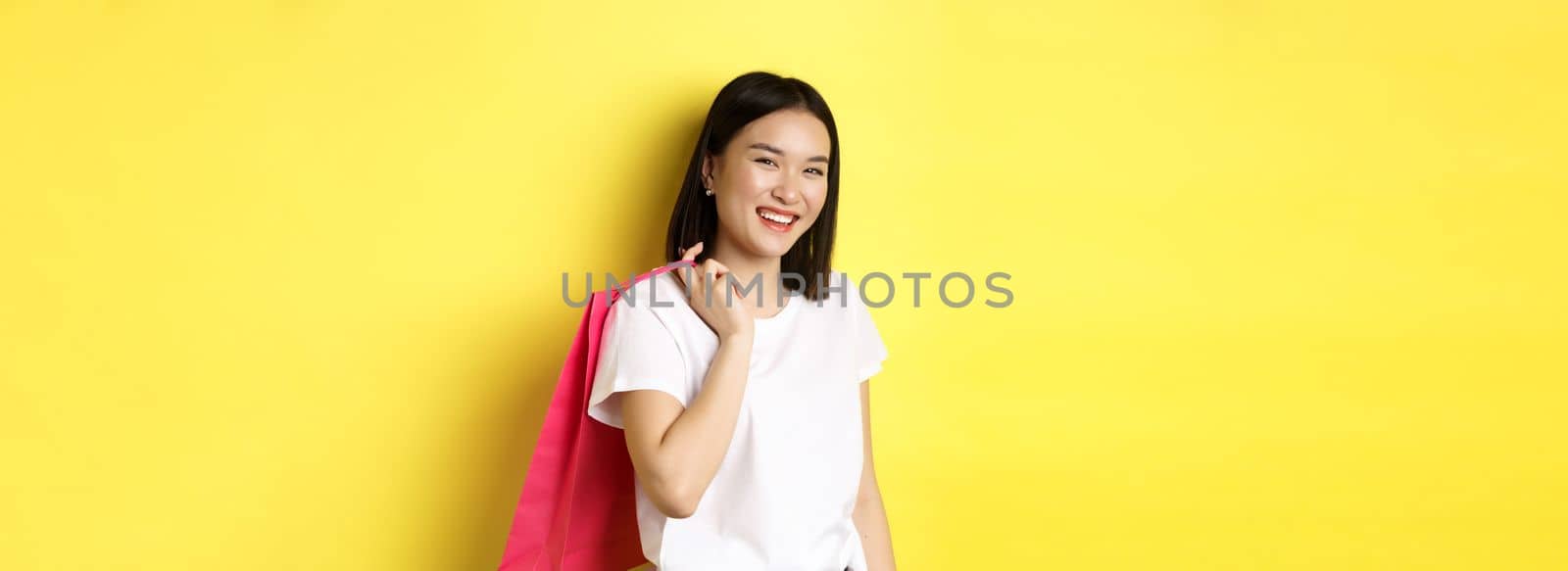 Happy asian woman holding shopping bag behind shoulder and laughing, standing over yellow background by Benzoix