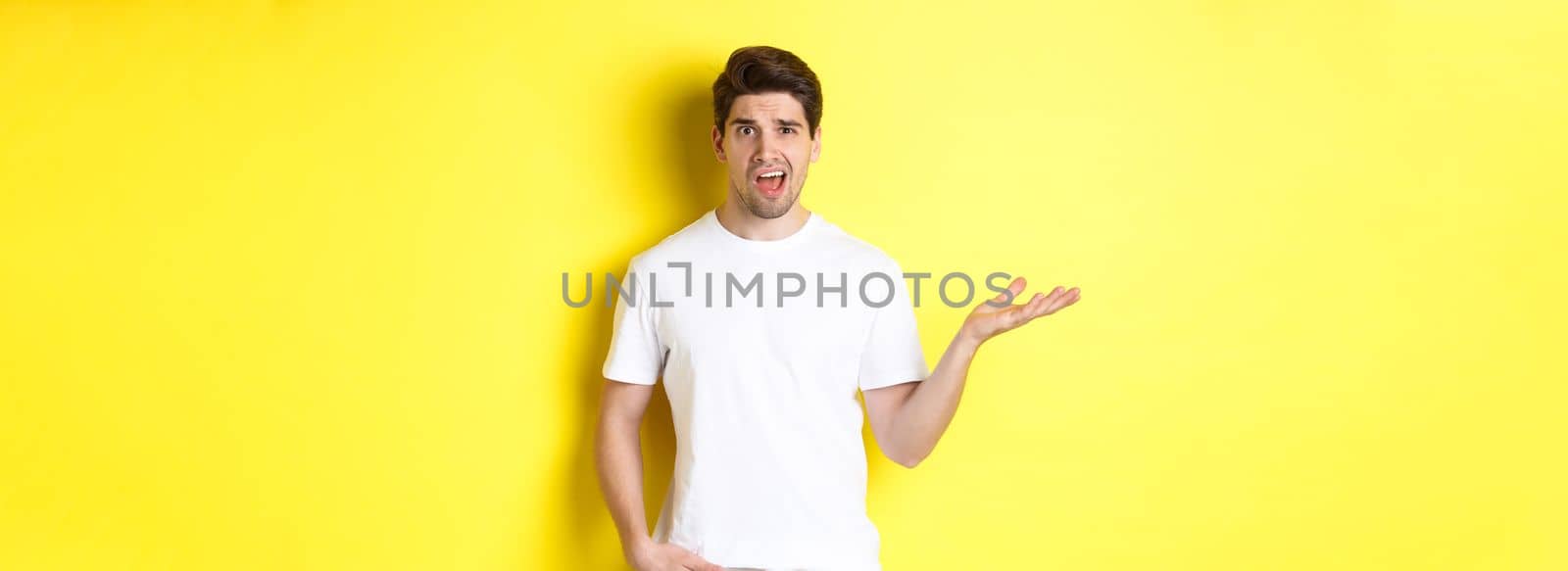 Confused and shocked man complaining, raising one hand and looking bothered, standing near yellow copy space.