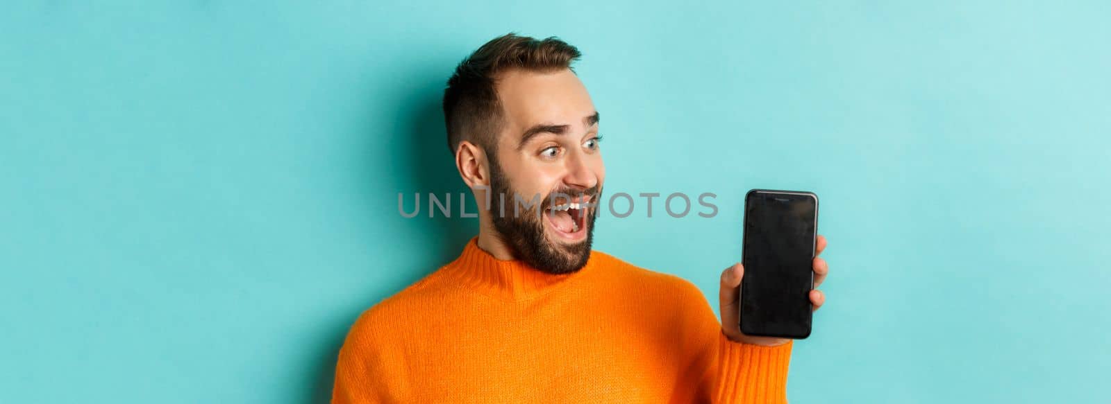 Close-up of young bearded man showing phone screen and looking amazed, wearing orange sweater, standing against studio background.