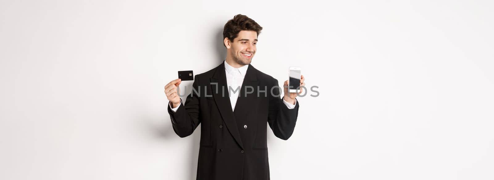 Handsome successful businessman, looking at smartphone screen and showing credit card, standing in black suit against white background.
