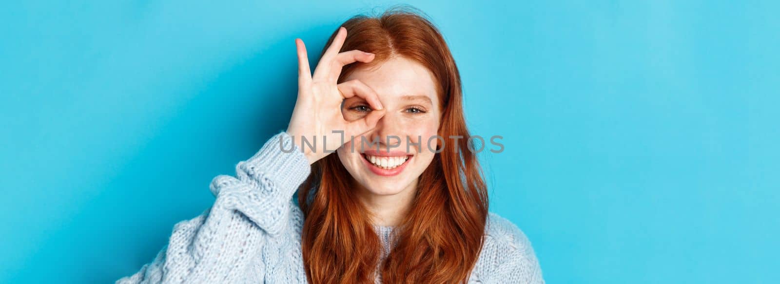Headshot of cheerful redhead female model showing okay sign over eye, smiling satisfied and happy, standing against blue background by Benzoix