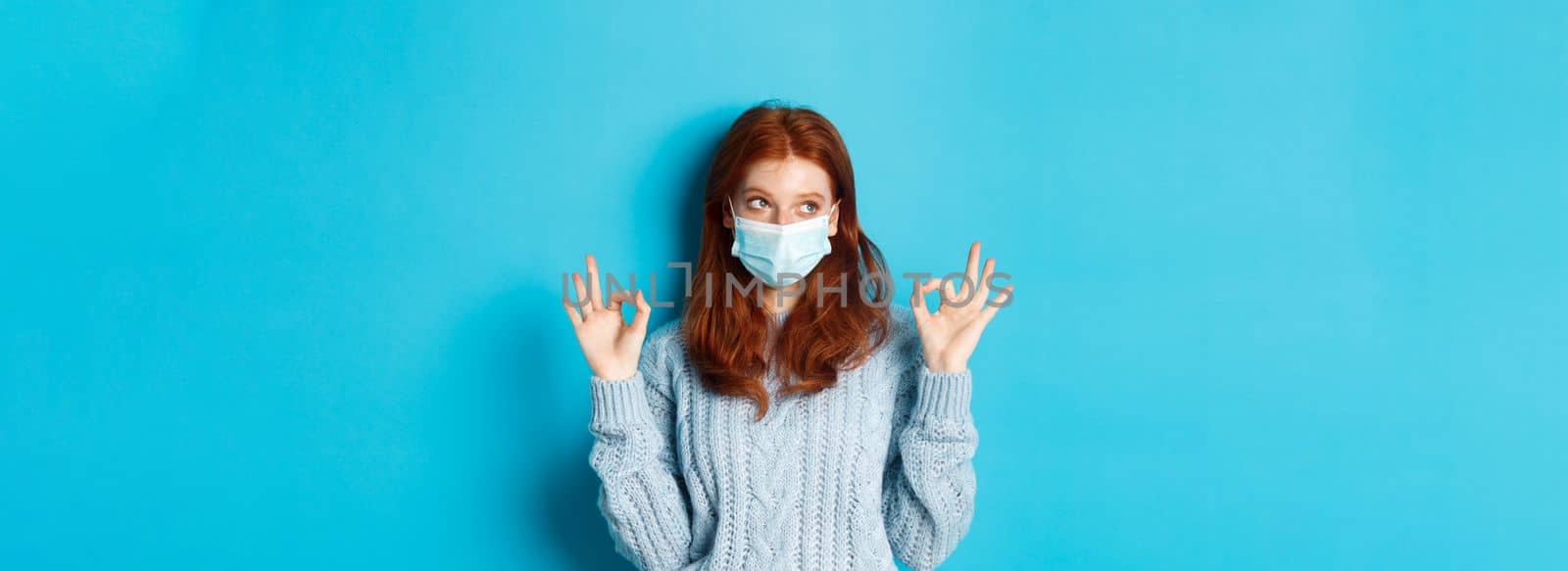 Winter, covid-19 and social distancing concept. Satisfied young redhead woman in face mask showing alright, okay gestures and looking left at promo, blue background.