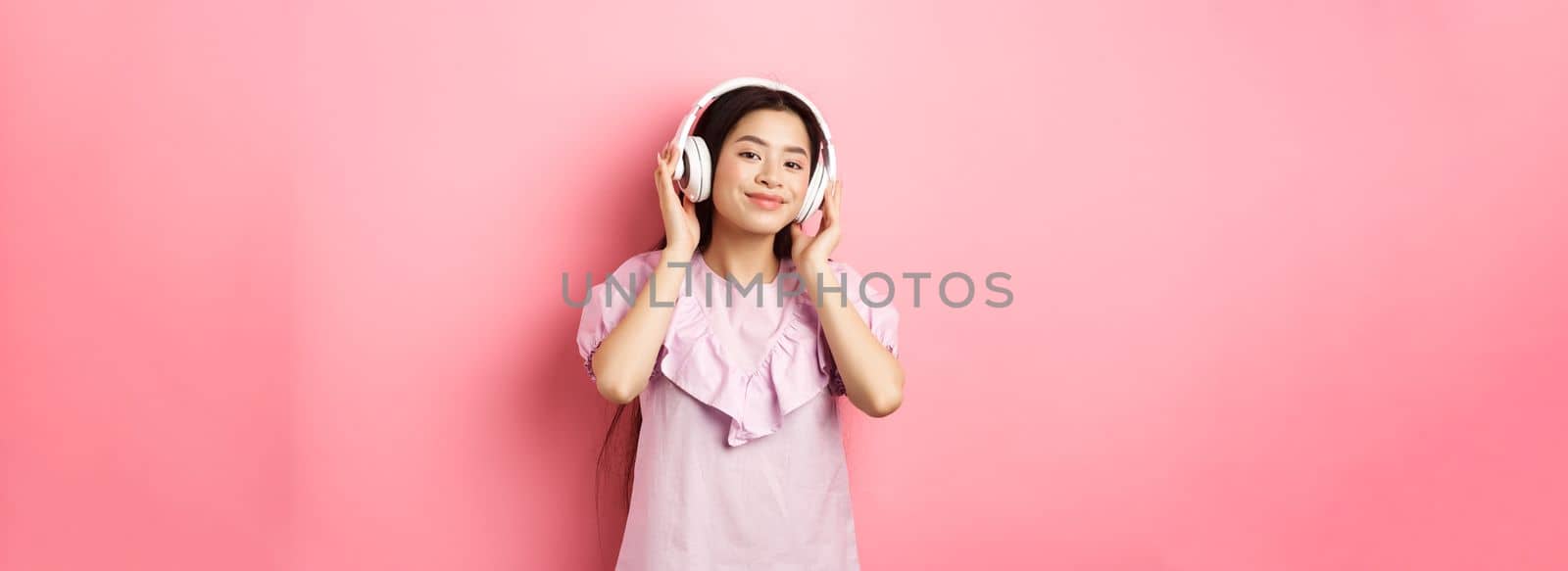 Beautiful asian woman listening music on wireless headphones, smiling pleased at camera, standing in dress against pink background by Benzoix