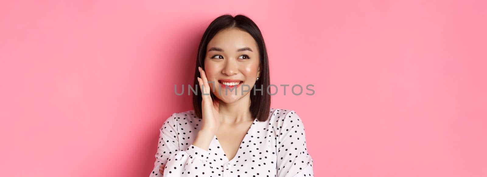 Coquettish asian woman in blushing, touching cheek and looking left amused, standing over pink background by Benzoix