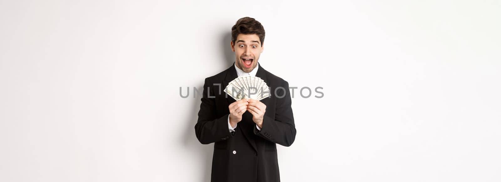 Image of handsome bearded guy in black suit, looking at money with excitement, standing over white background.