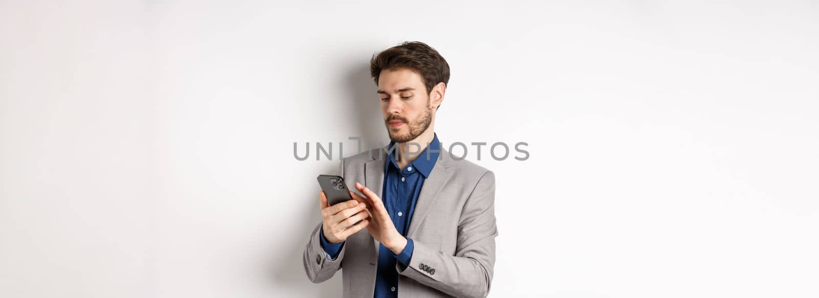 Young businessman in suit texting message on mobile phone, looking at smartphone, standing against white background.