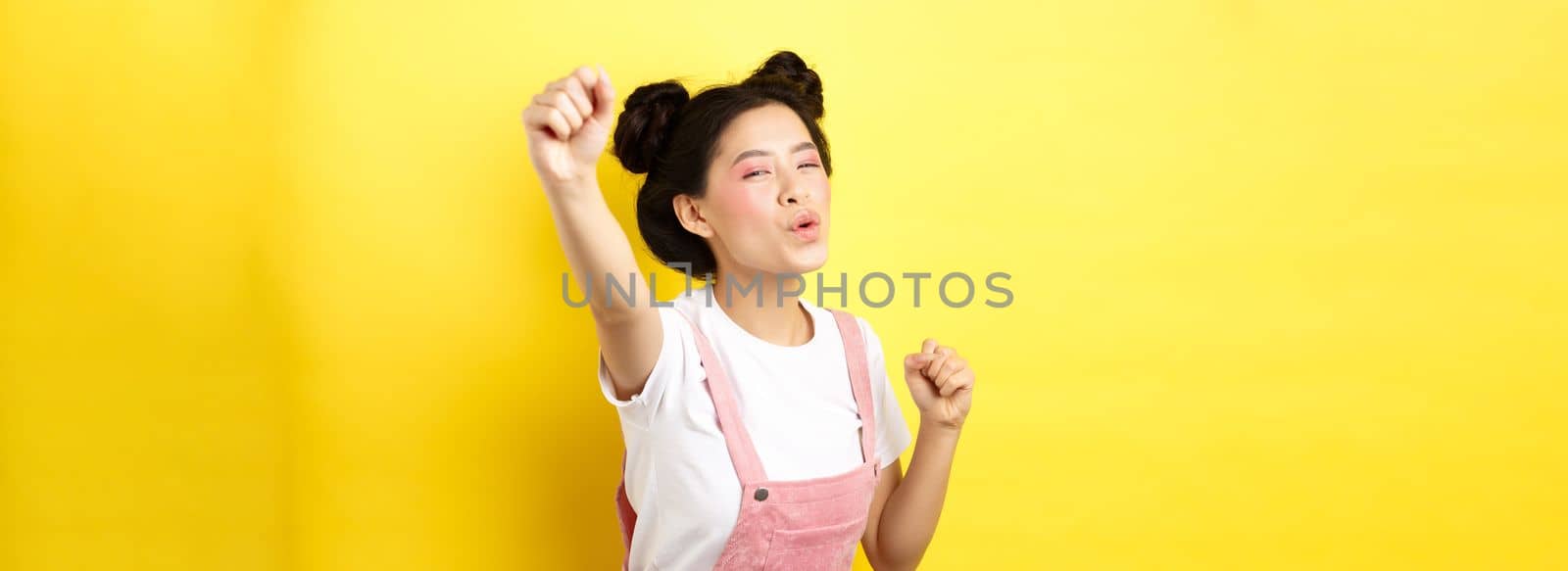 Excited asian girl looking motivated, raising hand up and chanting, cheering with joy, standing happy on yellow background.