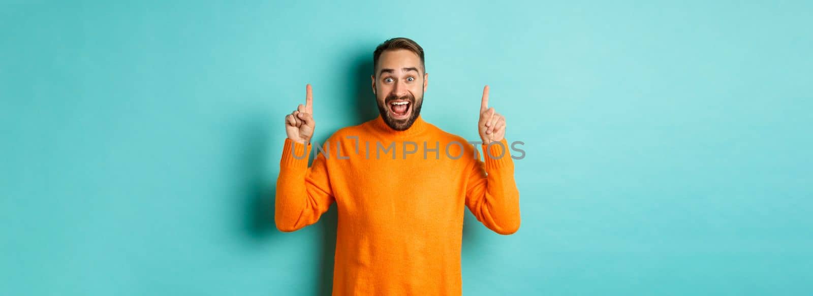 Young happy man showing advertisement, pointing fingers up announcing promo offer, standing over turquoise background by Benzoix