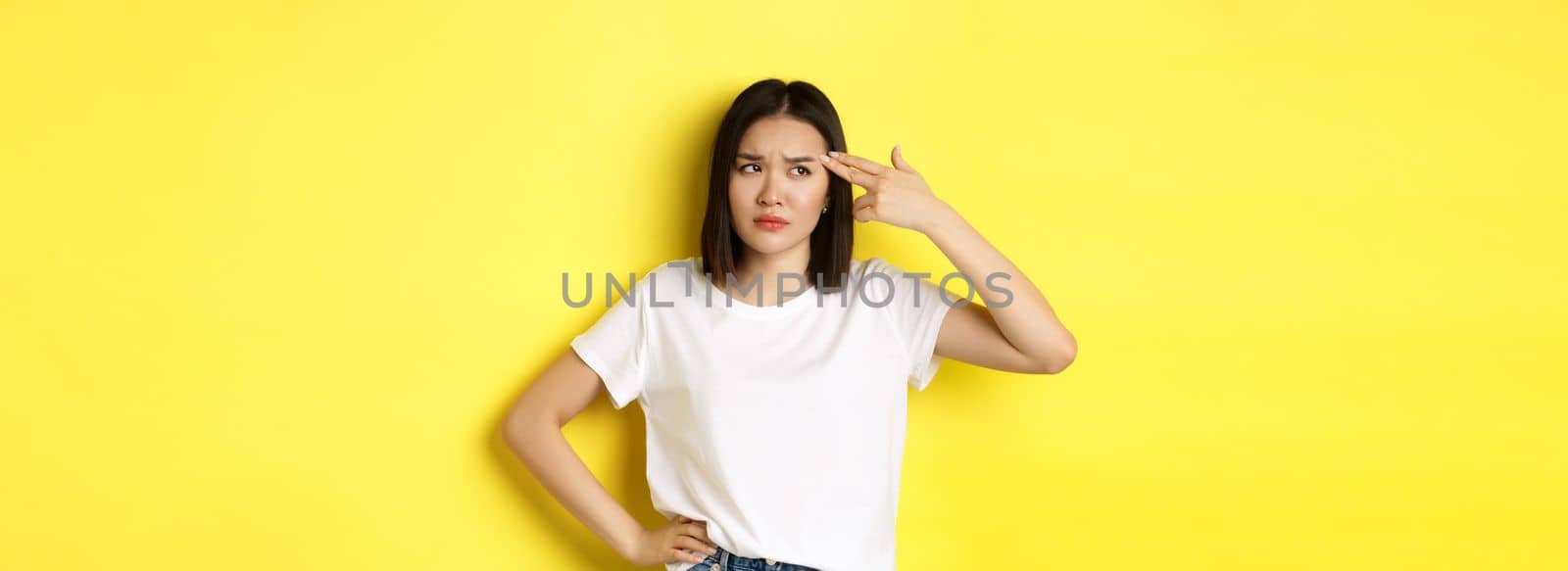 Woman showing kill me please gesture, shooting herself in head with finger gun from boredom, standing upset over yellow background.