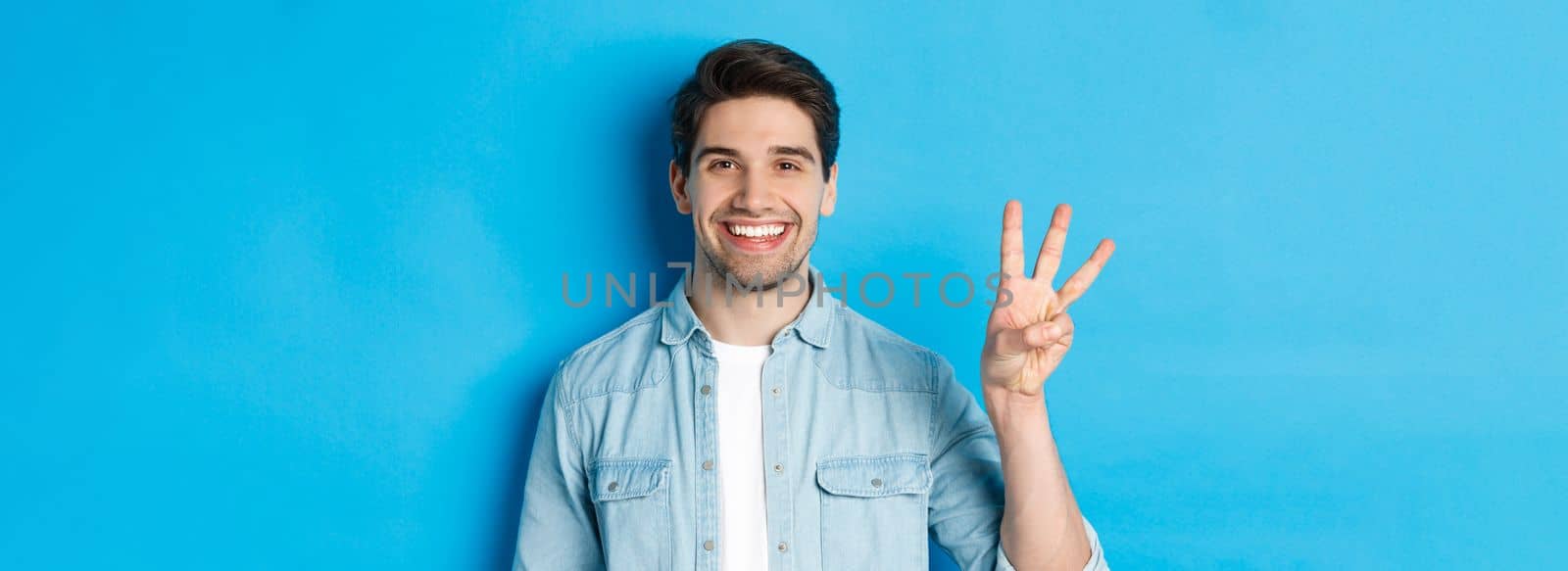 Close-up of handsome man smiling, showing fingers number three, standing over blue background.