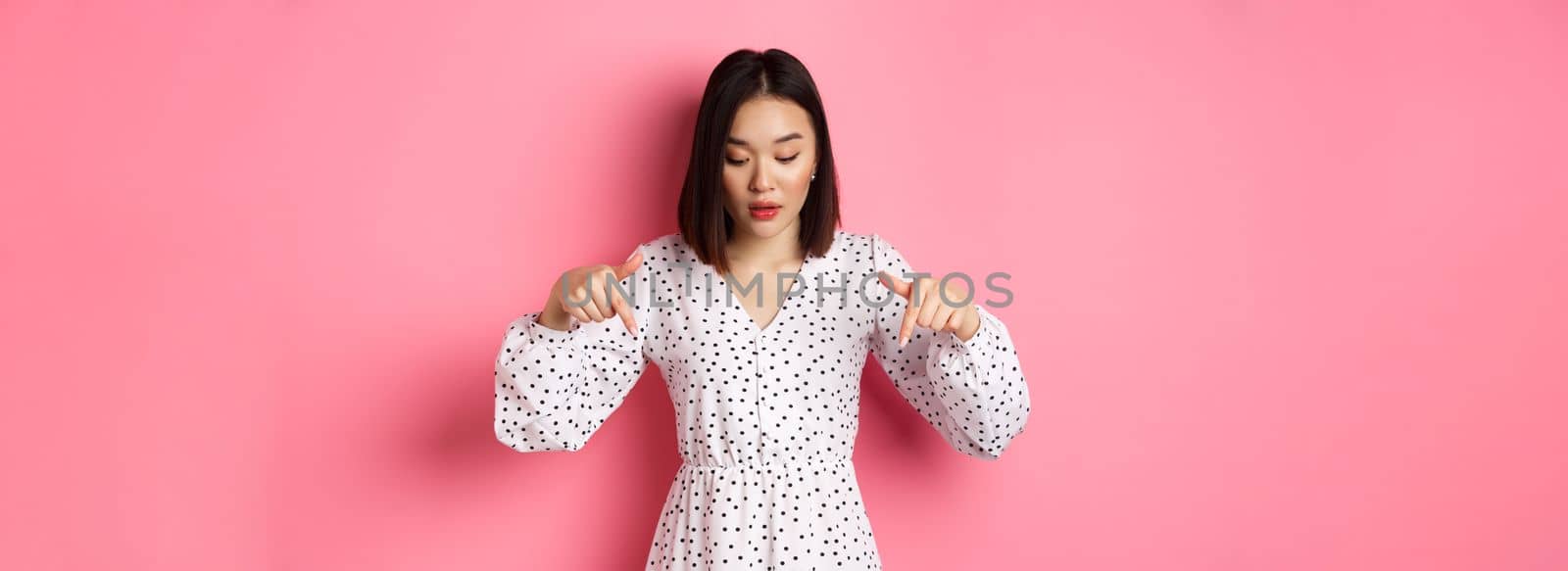 Romantic asian woman peeking down, pointing fingers at bottom, looking curious at product discount, standing over pink background.