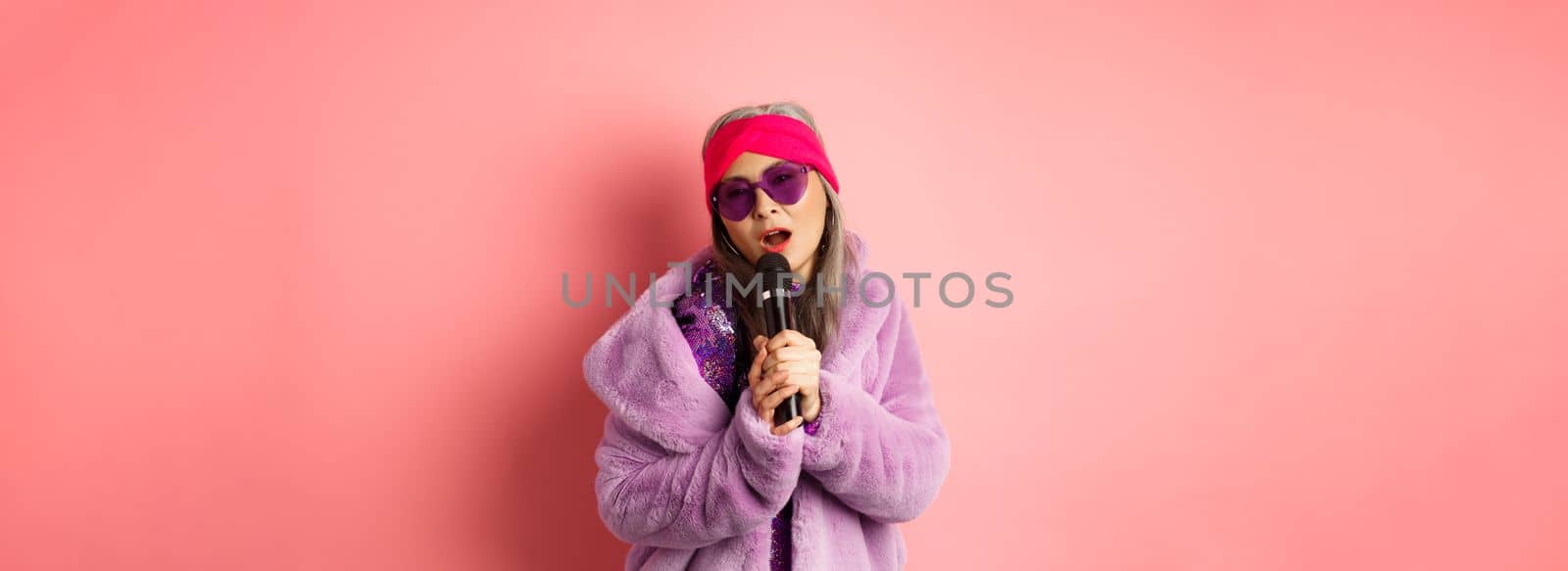 Fashion concept. Stylish senior asian woman singing karaoke, perform on stage with microphone, wearing trendy sunglasses and purple faux fur coat, pink background.