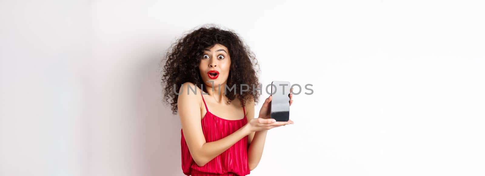 Excited woman with curly hair, makeup and red dress, gasping amazed, showing empty smartphone screen with amazed face, standing against white background by Benzoix