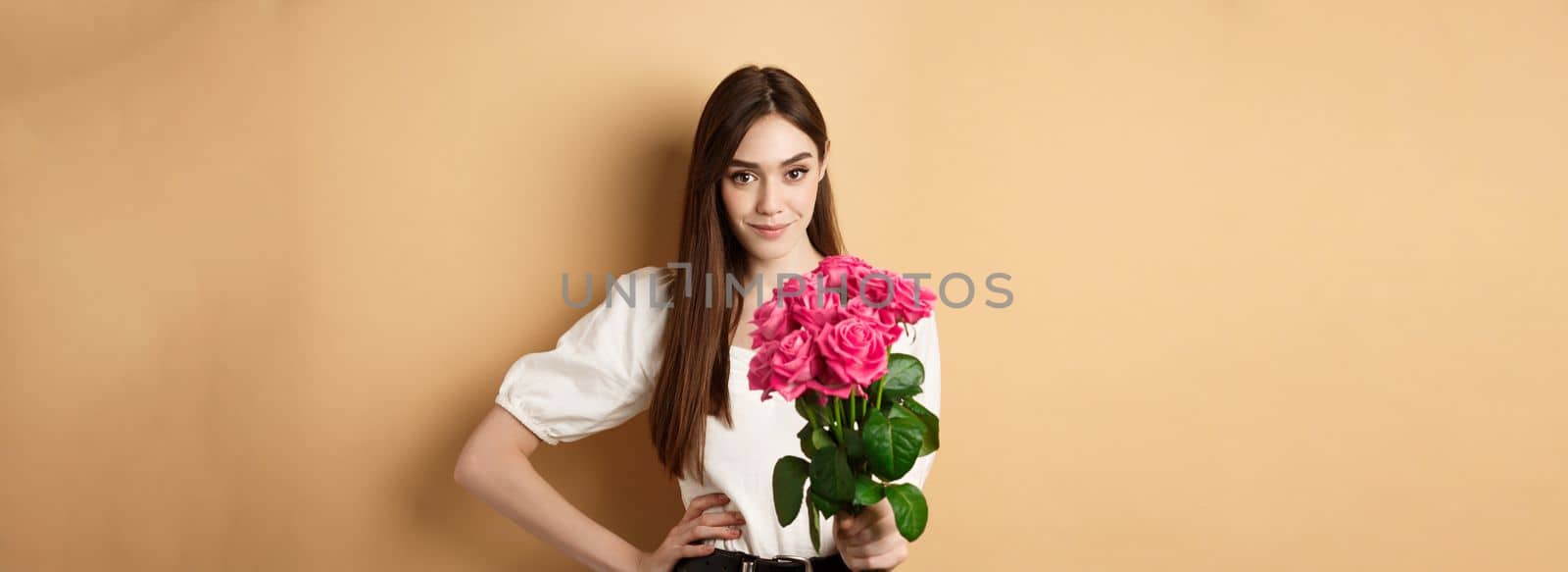 Valentines day. Beautiful girlfriend holding pink roses and looking at camera. Young woman receive flowers from her date, standing on beige background by Benzoix
