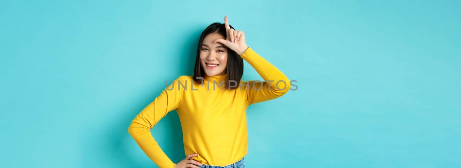 Sassy asian girl mocking lost team, showing loser sign on forehead and smiling pleased, being a winner, standing over blue background by Benzoix