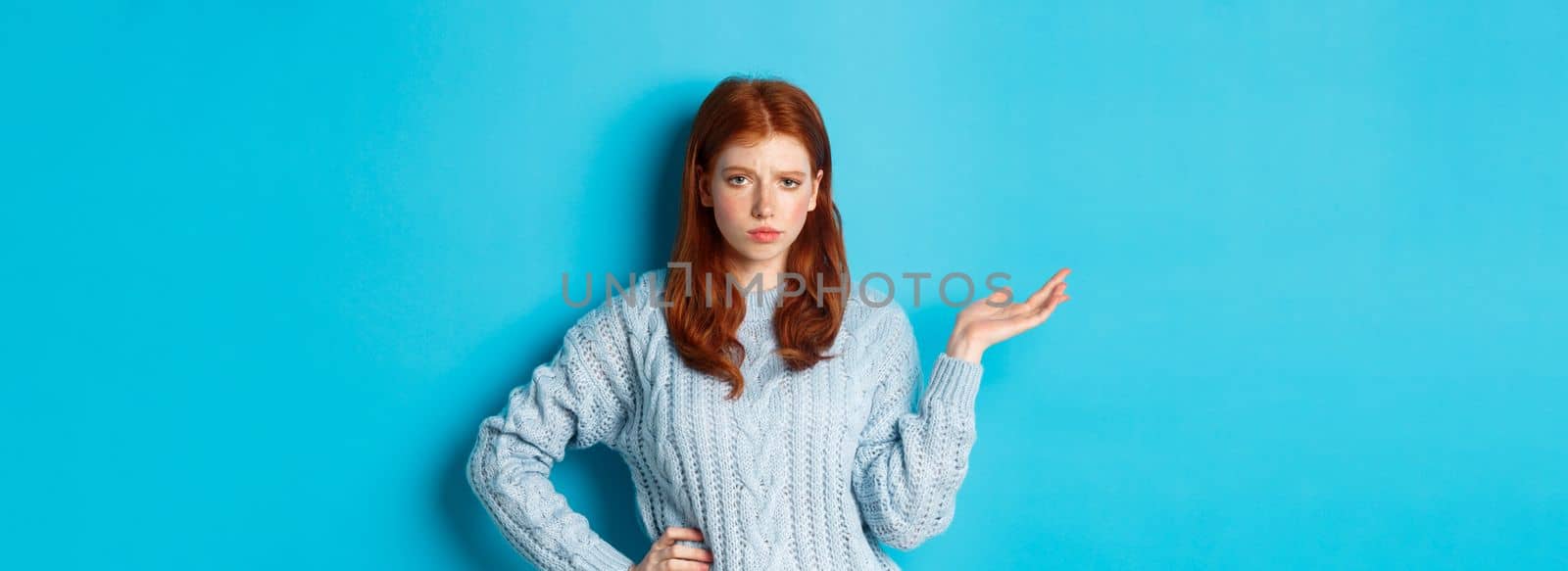 Skeptical teenage girl looking unamused, raising hand in so what gesture, staring at something with careless face, standing over blue background.