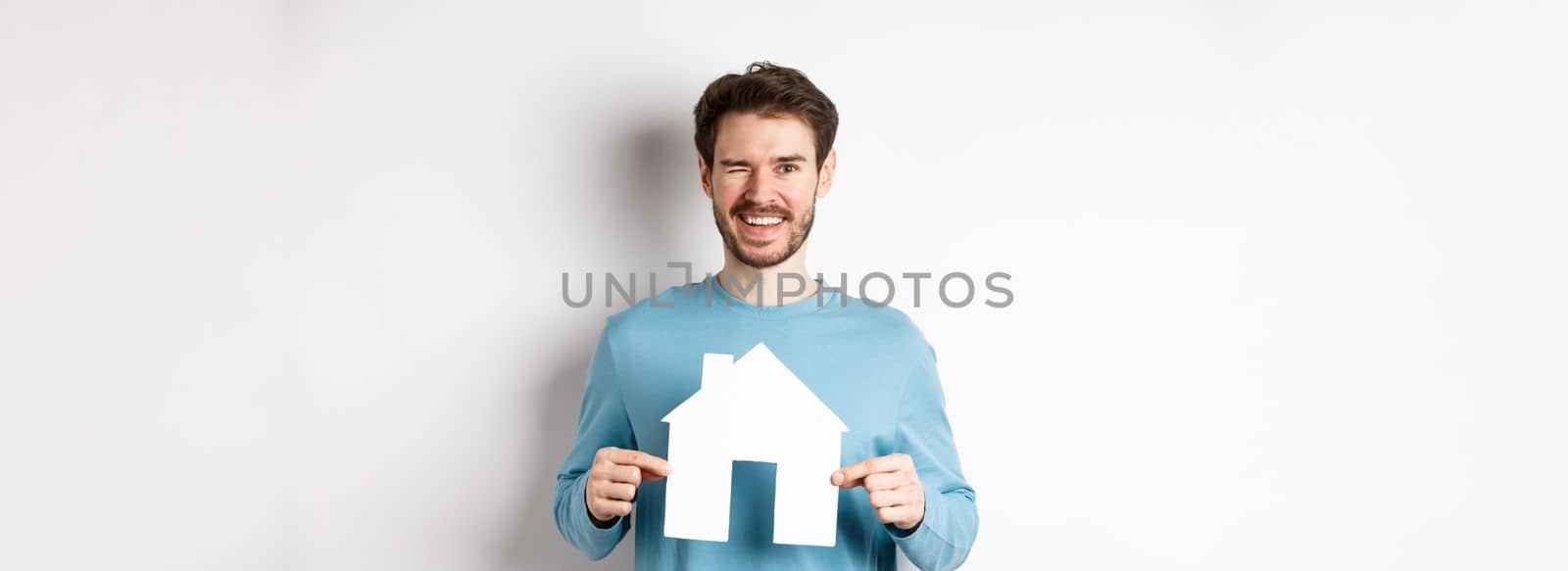 Real estate and insurance concept. Happy young man winking and smiling, showing paper house cutout, standing over white background by Benzoix