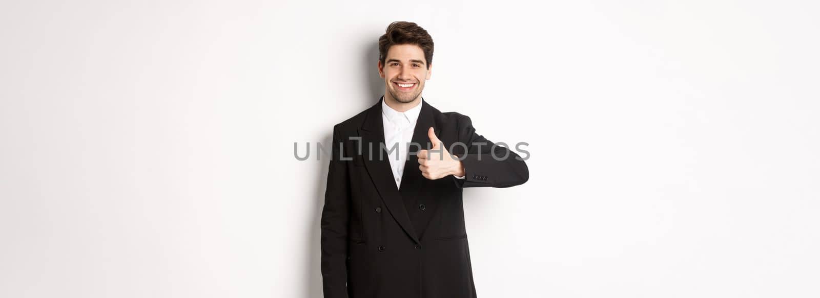 Portrait of handsome and confident male realtor, showing thumb-up and smiling, guarantee quality and recommending company, standing over white background.