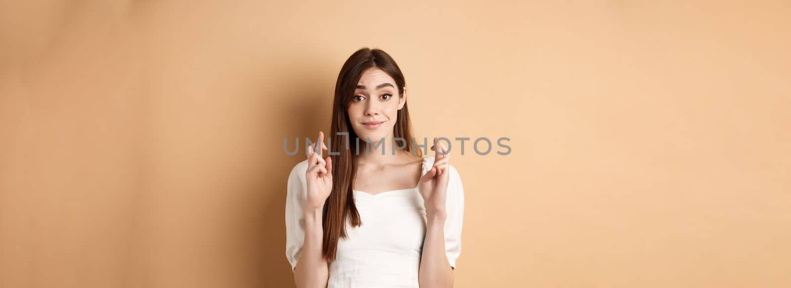 Hopeful tender girl in white blouse making wish, holding fingers crossed and smiling, praying for good luck, waiting for results, beige background by Benzoix