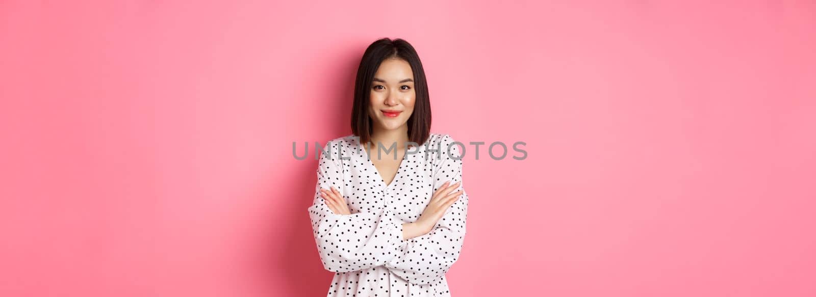 Beautiful asian female model standing in dress, cross arms on chest and smiling at camera, standing over pink background by Benzoix