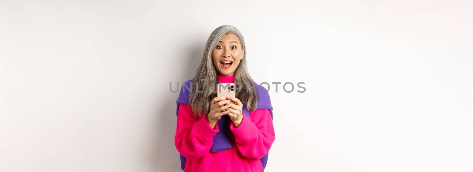 Surprised asian woman smiling at camera after reading promotion on smartphone, standing with mobile phone over white background.