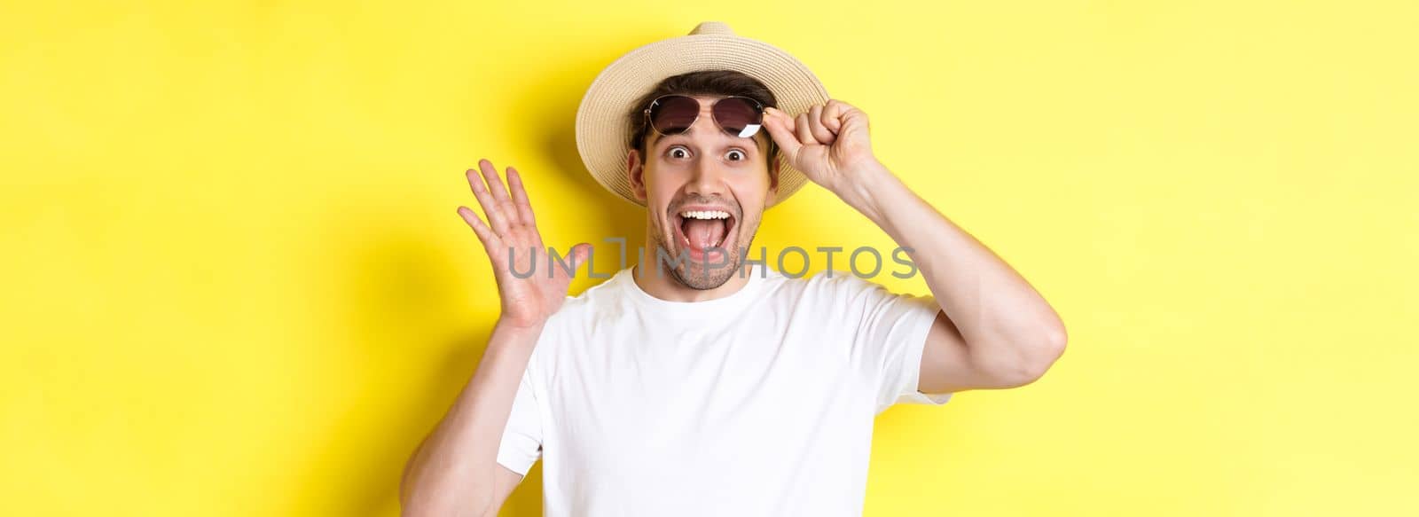 Concept of tourism and holidays. Excited man tourist take-off sunglasses and looking amazed at promo offer, standing over yellow background by Benzoix
