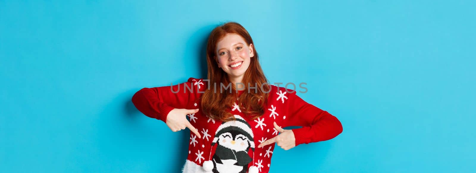 Winter holidays and Christmas Eve concept. Pretty redhead girl pointing fingers at cute xmas sweater with penguin, standing over blue background.