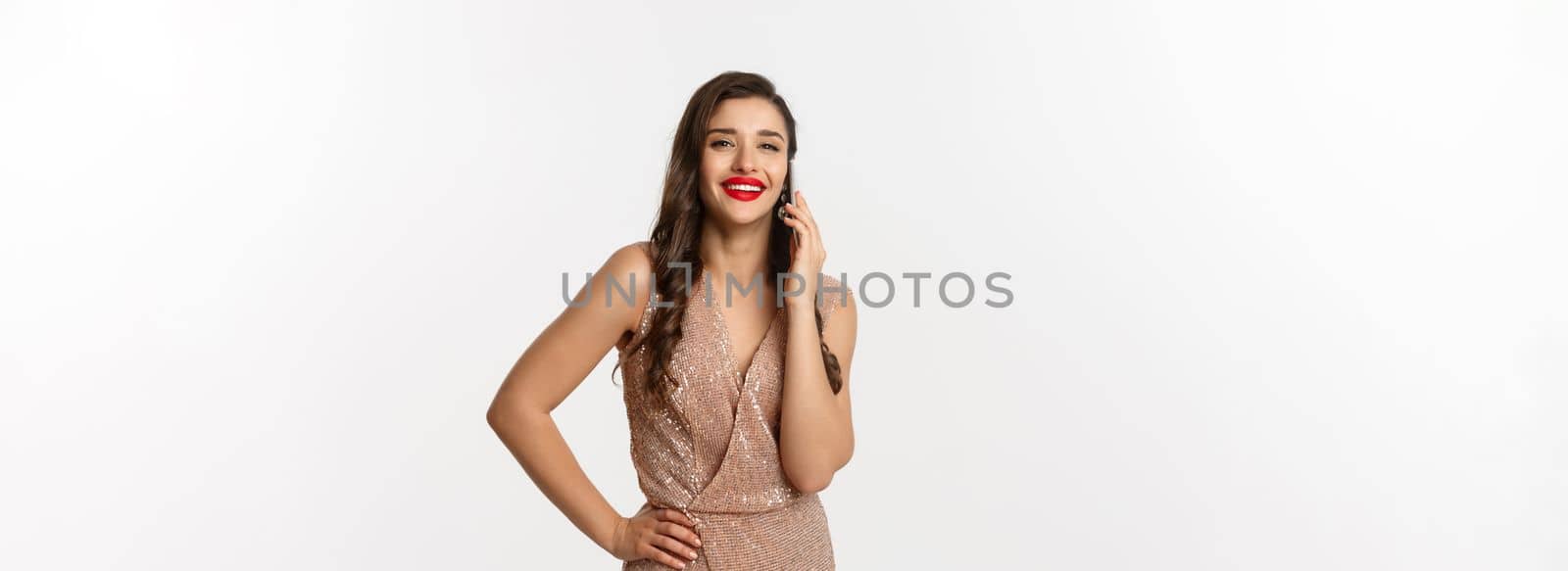 Christmas party and celebration concept. Attractive young woman with red lips, wearing luxury dress, talking on mobile phone and smiling, standing over white background.