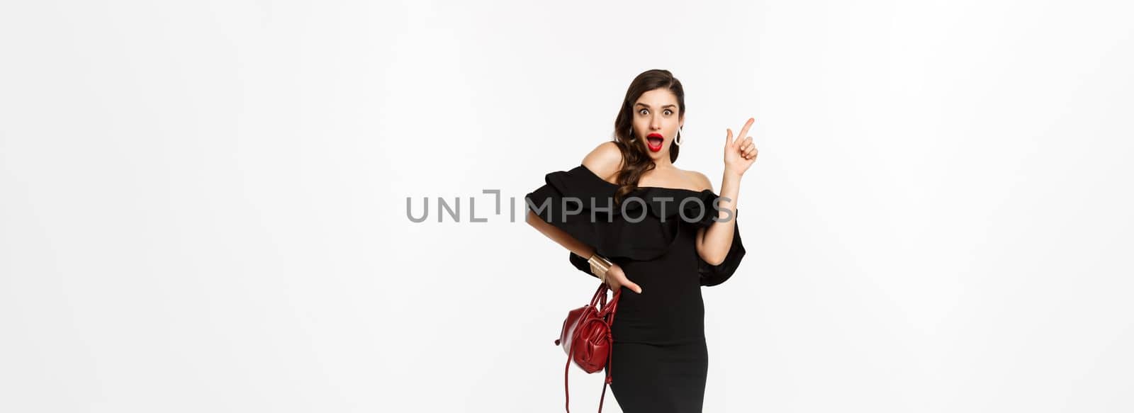 Beauty and fashion concept. Full length of excited young woman in glamour dress, red lips, having an idea, raising finger to suggest something, white background.