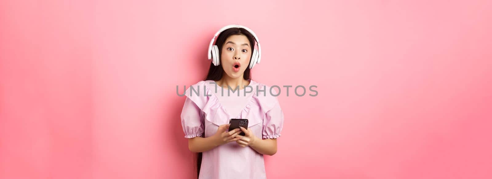 Excited asian woman in wireless headphones say wow, holding mobile phone and looking amused at camera, standing against pink background by Benzoix
