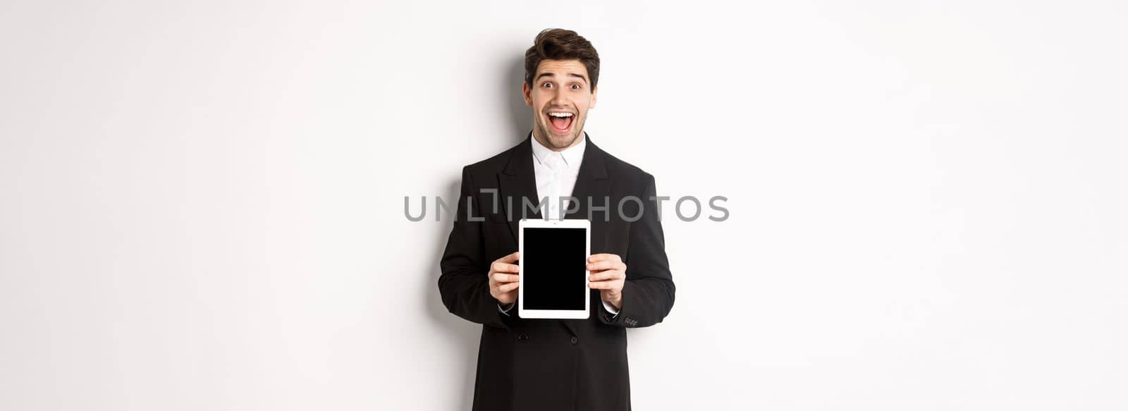 Image of attractive male entrepreneur in trendy suit, showing digital tablet screen and smiling amazed, standing over white background.