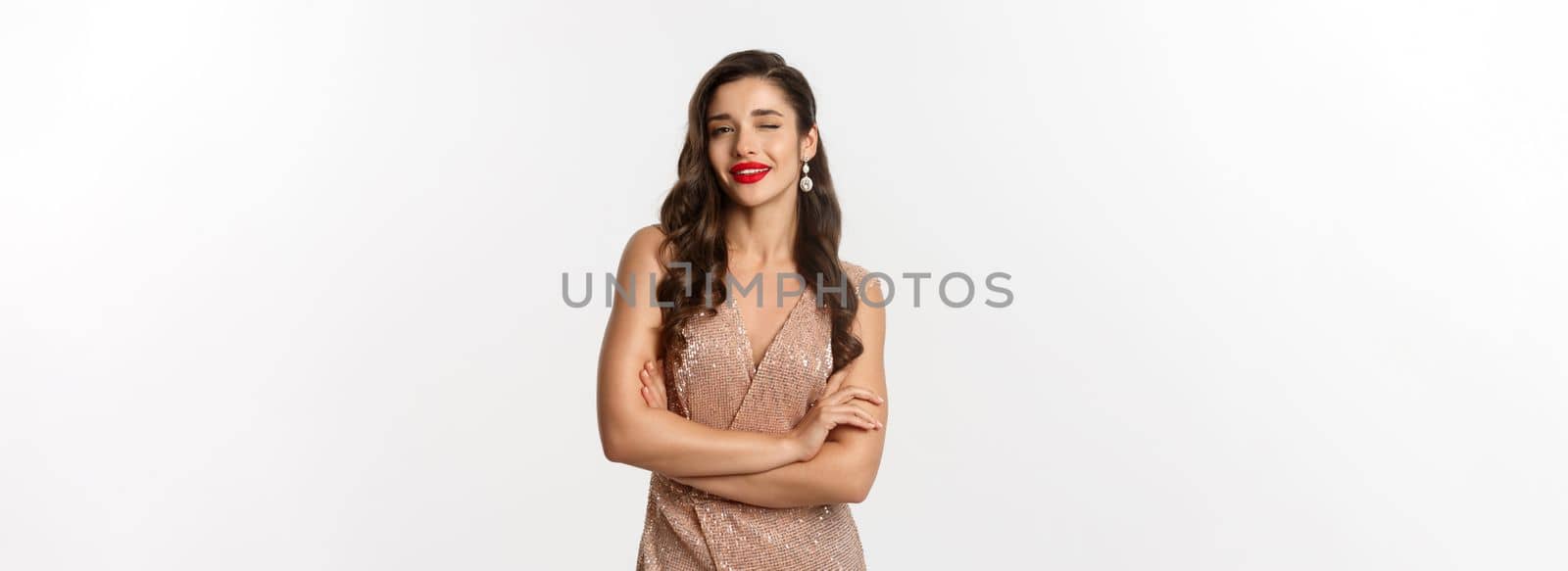 Beautiful woman in evening dress and red lipstick, winking at camera and smiling, celebrating New Year party, standing over white background.