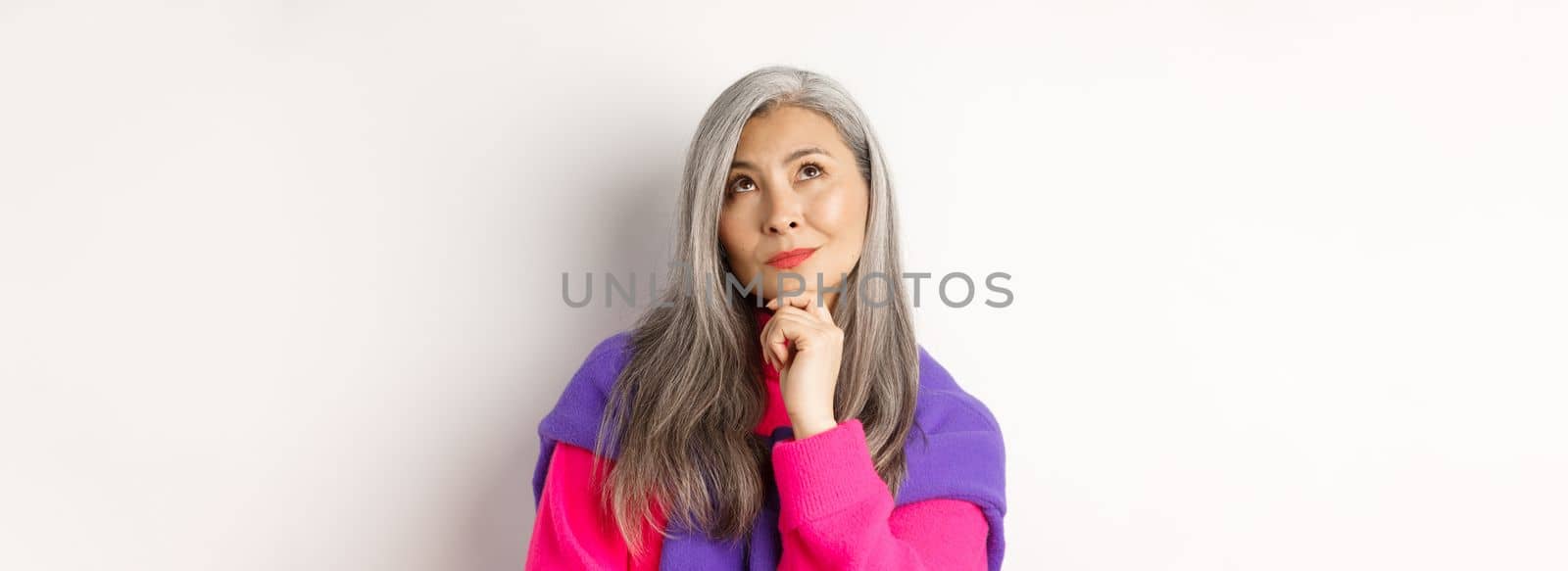 Close up of thoughtful asian female with grey hair, looking up and thinking, making decision, standing over white background.