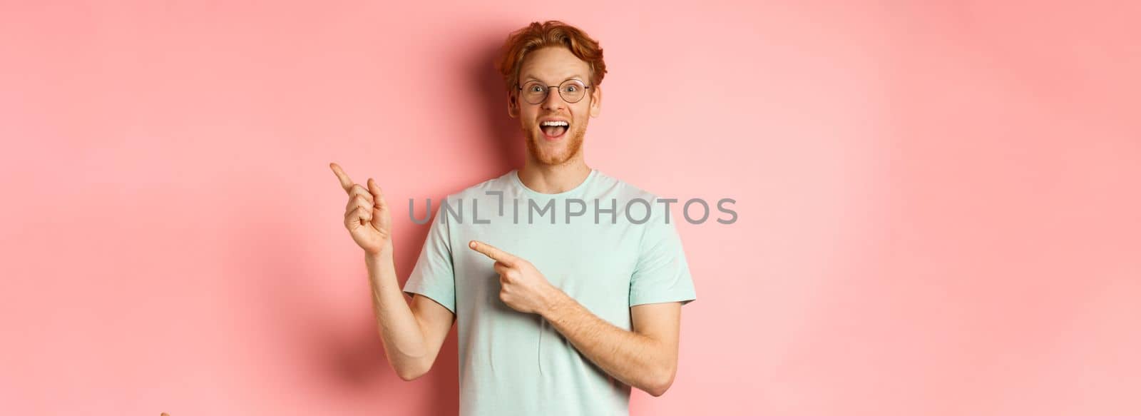 Amazed redhead man in glasses and t-shirt showing special promotion, pointing fingers at upper right corner and staring excited at camera, standing over pink background by Benzoix