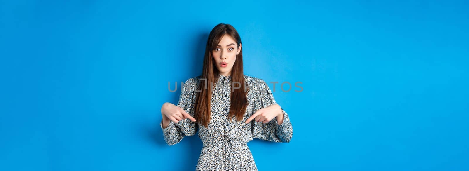 Excited beautiful woman in dress say wow, pointing fingers down and look amused, checking out promotion, standing on blue background.