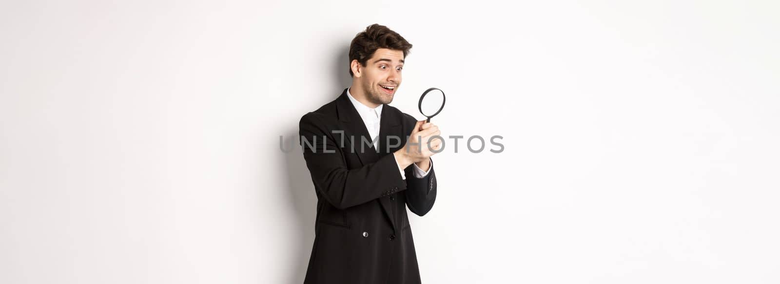 Image of handsome businessman in black suit, looking through magnifying glass, found something and smiling, standing over white background.