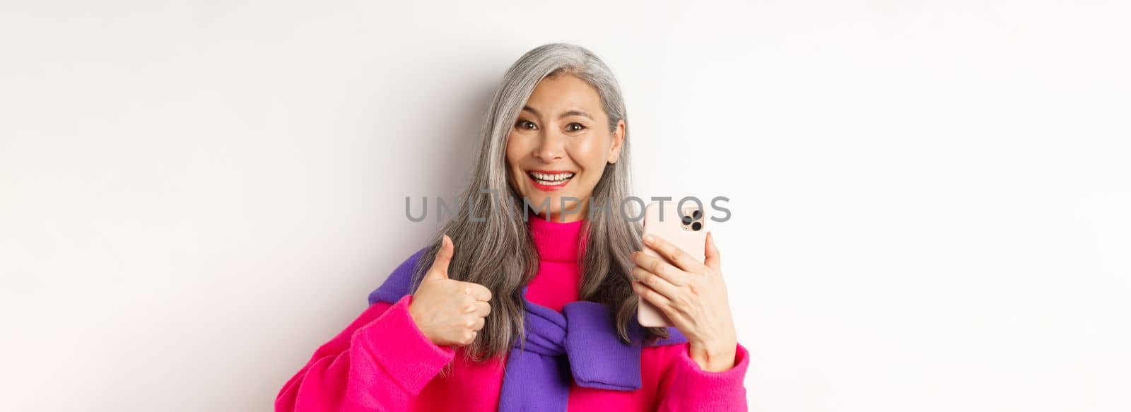 Online shopping. Close up of fashionable asian senior woman showing thumbs-up, using smartphone and approve something, standing over white background.