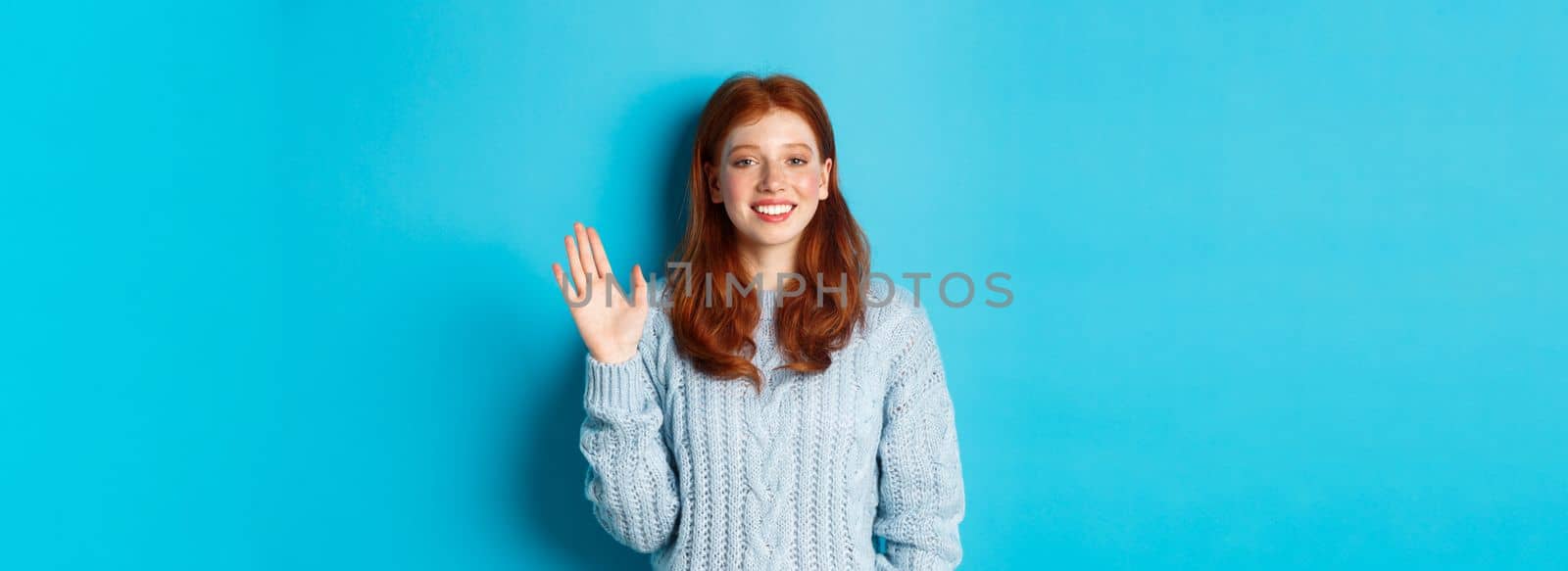 Friendly redhead teenage girl saying hi, waving hand in hello gesture and smiling, standing against blue background.