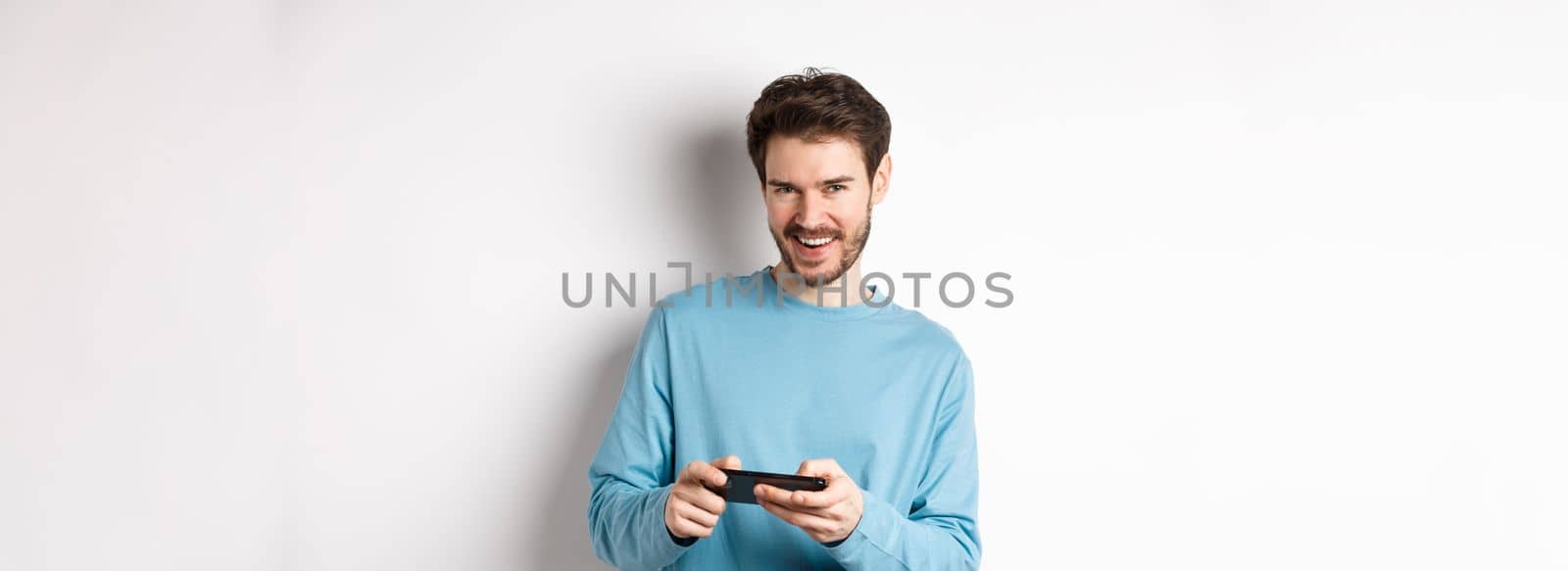 Image of handsome young man playing mobile video game, holding smartphone horizontally and smiling at camera pleased, standing over white background.