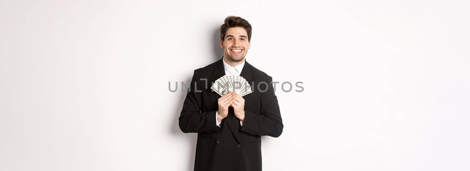 Portrait of happy and pleased handsome man in suit, hugging money and looking satisfied, standing over white background.