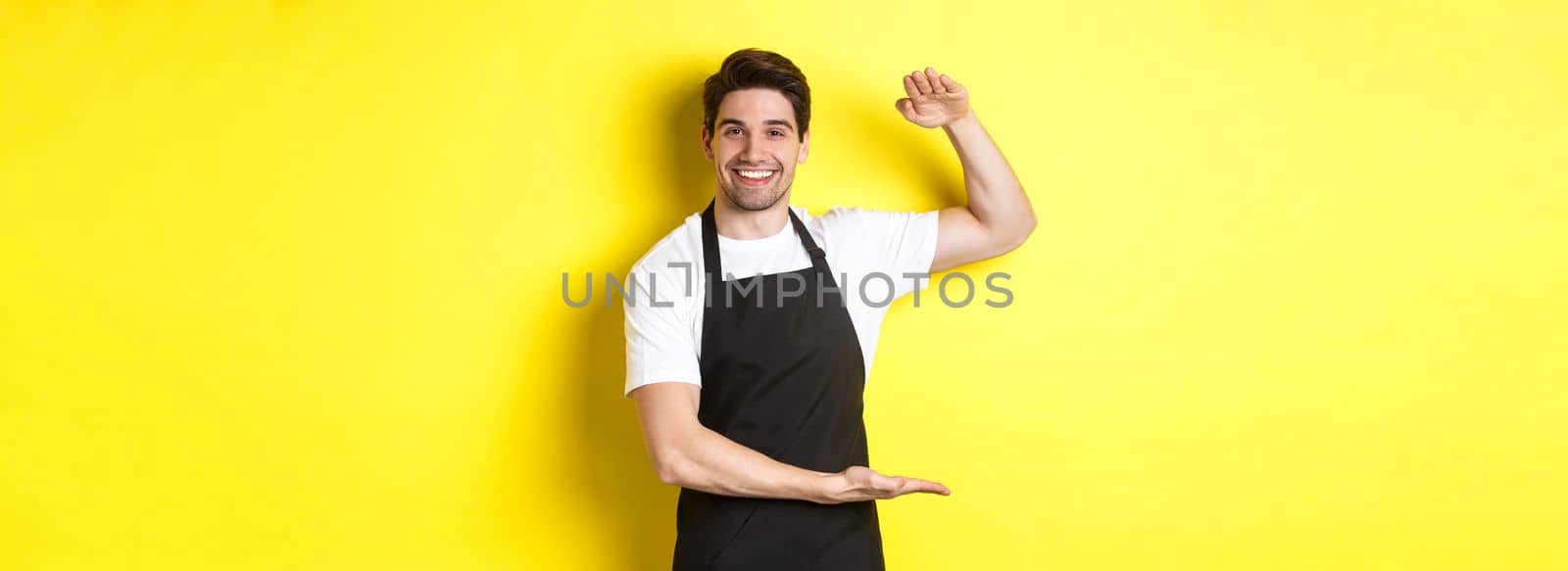 Smiling handsome barista showing something long or large, standing over yellow background.