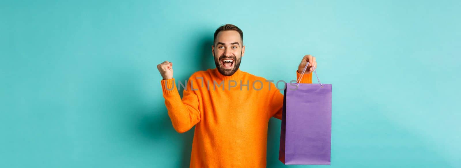 Happy man holding purple shopping bag and rejoicing, got discount and celebrating, standing over turquoise background by Benzoix