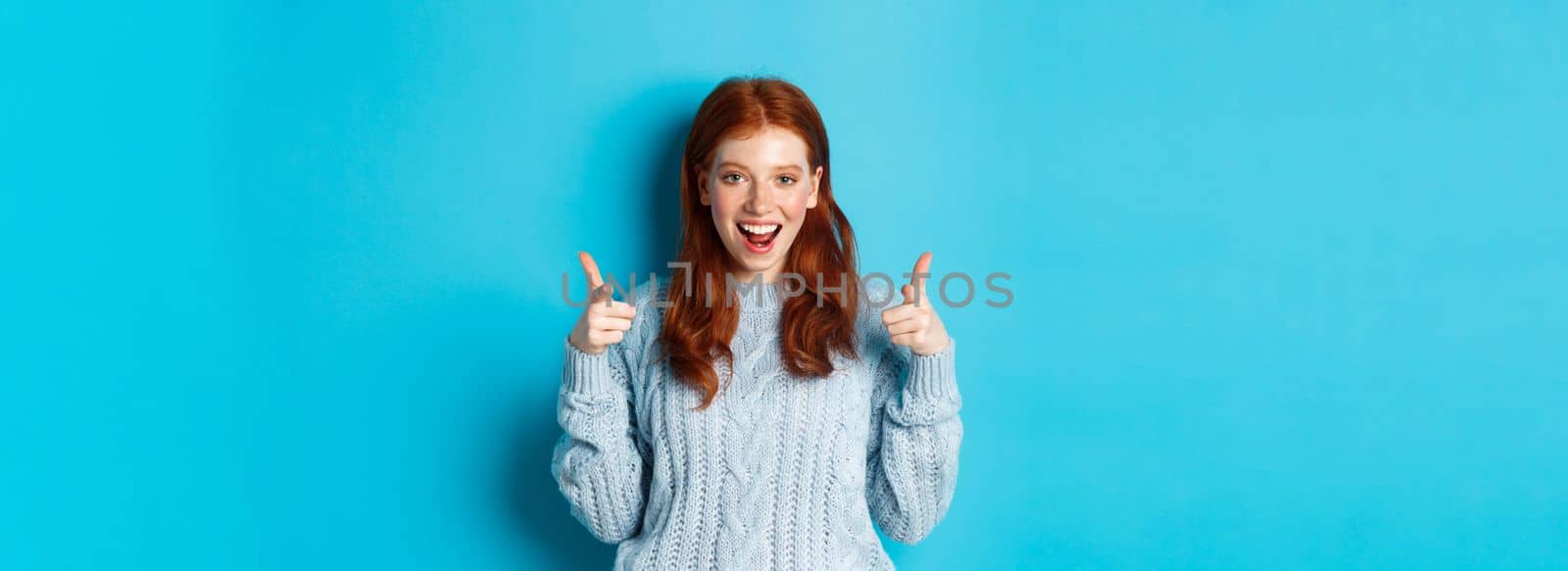 Cheerful teenage girl showing approval, make thumbs up and agree, liking something, standing over blue background.