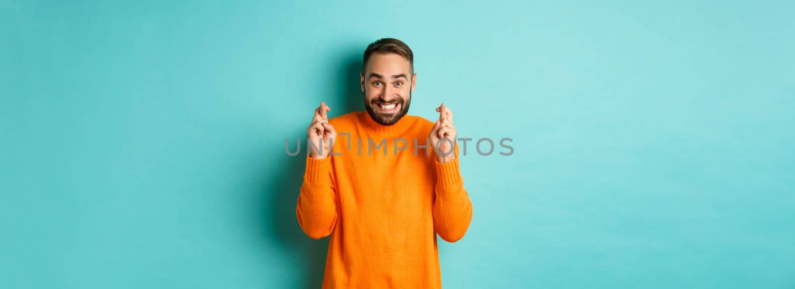 Excited man with beard, making a wish, holding fingers crossed for good luck and smiling, standing over light blue background.