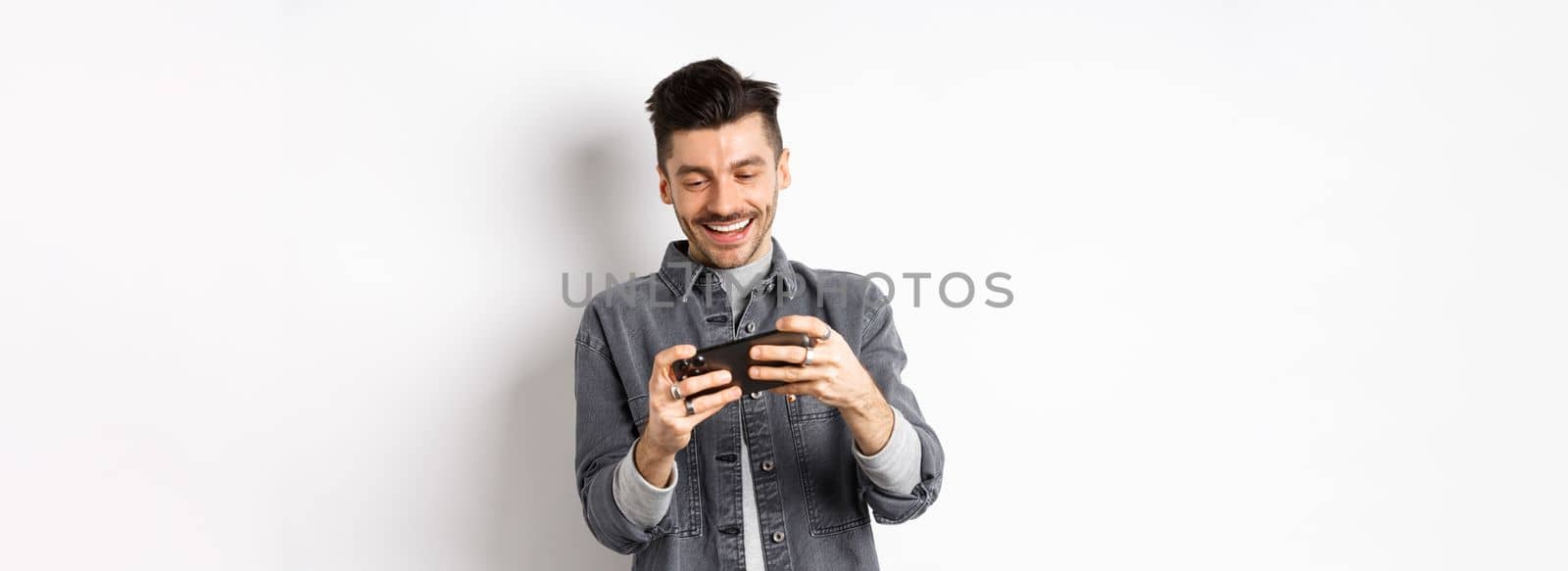 Handsome young man playing mobile video game and smiling at screen, standing against white background by Benzoix