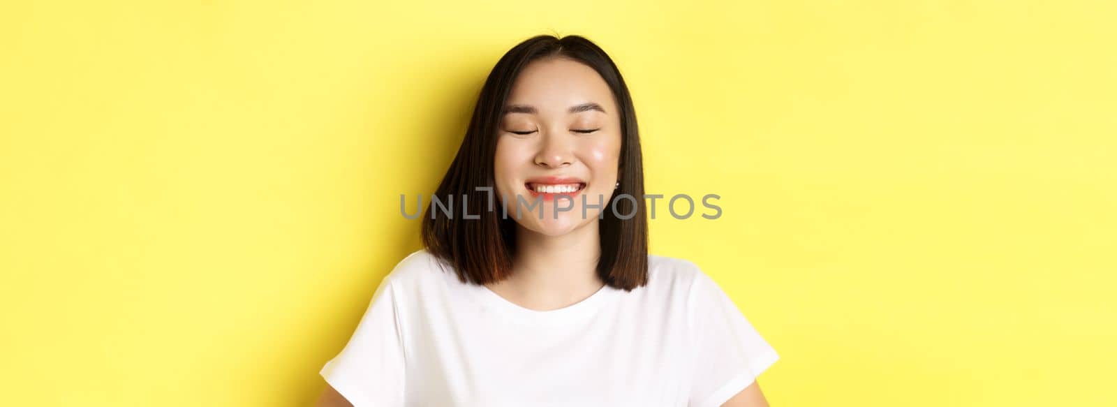 Close up of happy, romantic asian girl dreaming of something, close eyes and smiling delighted, standing over yellow background.