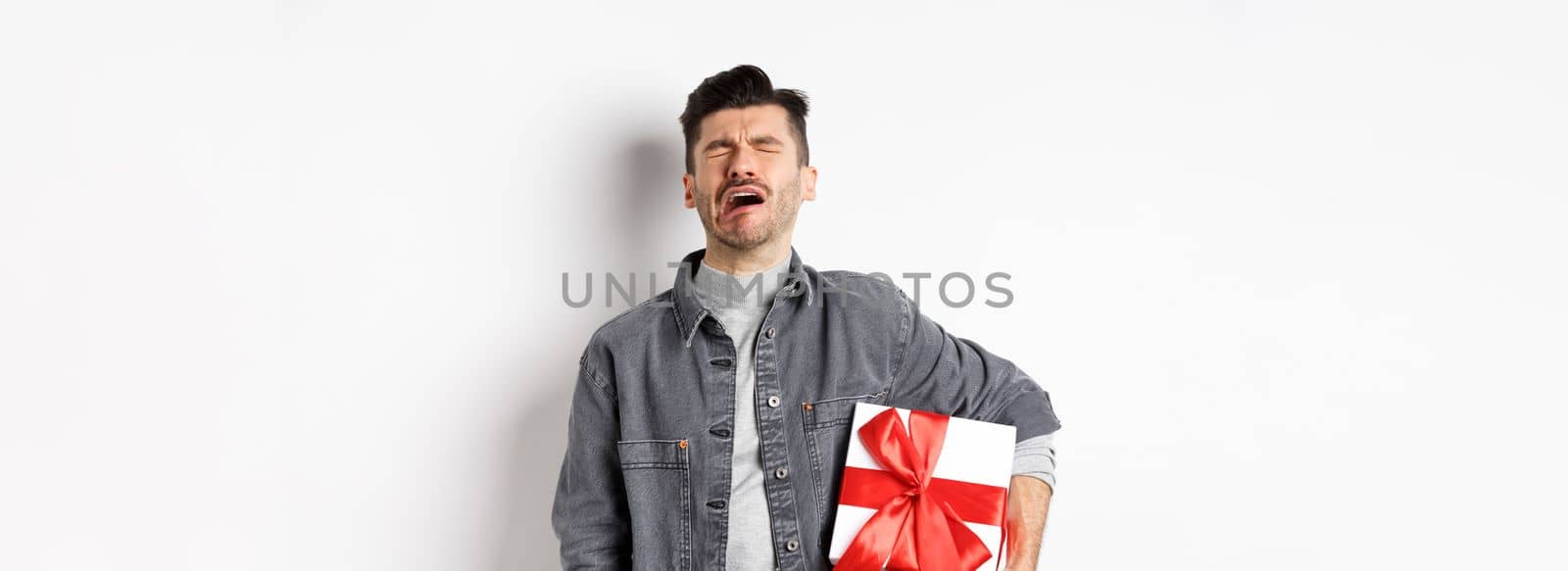 Valentines day heartbreak. Single and heartbroken guy crying lonely, holding big gift box, being rejected by lover, sobbing and feeling alone, standing on white background.